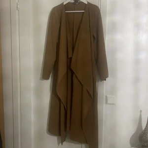 Brun kappa från Boohoo - Säljer en snygg brun kappa från Boohoo i storlek EUR 38. Den har en lång och elegant design med långa ärmar och en öppen framsida. Perfekt för höst och vår, och ger en stilren look till vilken outfit som helst. Passar både till vardags och festliga tillfällen.