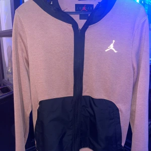 Grå och svart hoodie från Jordan - Säljer en snygg grå och svart hoodie från Jordan. Den har en dragkedja framtill och det ikoniska Jordan-loggan på bröstet. Perfekt för både vardag och träning. Hoodien har långa ärmar och en bekväm passform. Passar perfekt till höst och vår!
