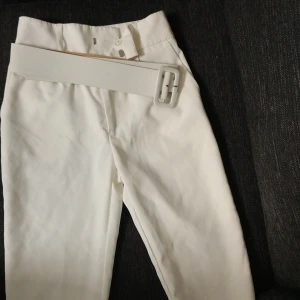 Vita byxor med bälte - Säljer ett par stilrena vita byxor i mycket bra skick. De är högmidjade och kommer med ett matchande bälte som ger en snygg detalj. Perfekta för både vardag och festliga tillfällen. Passar bra till vår och sommar!