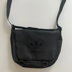 Adidas axelremsväska  - Säljer en snygg svart axelväska från Adidas i bra skick. Väskan har en justerbar axelrem och ett stort fack med dragkedja på baksidan. Perfekt som skolväska eller som daglig väska. Adidasloggan har små defekter som syns på när håll.