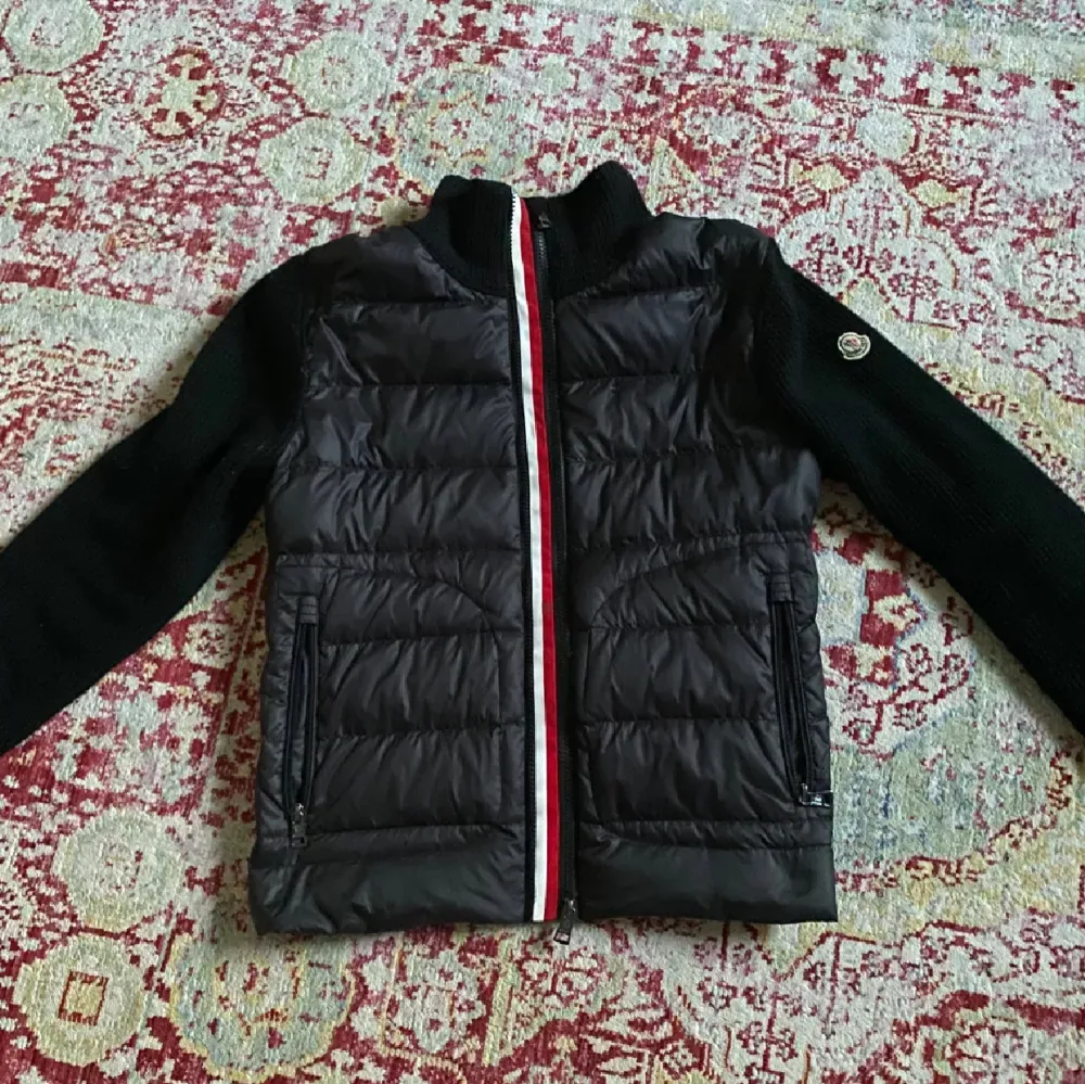 Säljer en snygg svart jacka från Moncler. Den har en quiltad design med dragkedja i rött, vitt och blått. Ärmarna är stickade och långärmade, vilket gör den perfekt för höst och vinter. Jackan är i mycket bra skick och passar både till vardags och lite finare tillfällen. OBS! ett litet hål vid bröstet dock syns den inte.. Jackor.