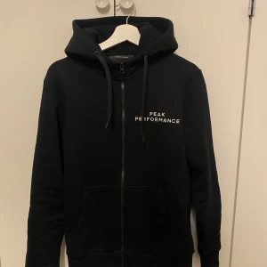 Svart hoodie från Peak Performance - Säljer en snygg svart hoodie från Peak Performance i mycket bra skick. Den har dragkedja framtill och en praktisk huva med snören. Perfekt för både vardag och träning. Stor logga på bröstet och en mindre på ärmen. Superbekväm och stilren!
