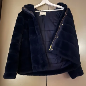 Mörkblå pälsjacka med huva - Säljer en supermjuk och varm mörkblå pälsjacka från samsoe samsoe. Den har en praktisk huva och stängs med dragkedja och knappar. Perfekt för kalla dagar när du vill vara både stilren och bekväm. Jackan är i mycket bra skick och passar perfekt till höst och vinter.
