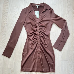 Brun klänning med knappar - Snygg brun klänning i mycket bra skick med långa ärmar och knappar hela vägen ner. Den har en smickrande rynkad detalj framtill som ger en fin passform. Perfekt för höst och vår!