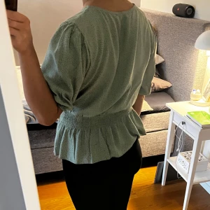 Grön blus med volangdetaljer - Säljer en söt grön blus med små prickar och volangdetaljer i midjan. Blusen har korta ärmar och en lös passform, perfekt för vår och sommar. Den är både bekväm och stilren. Använd en gång. 