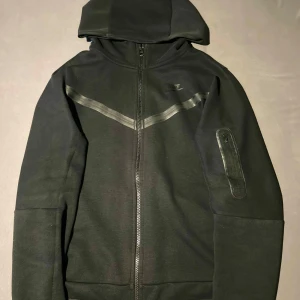 Svart hoodie från Nike - Säljer en svart hoodie från Nike i mycket bra skick. Den har en dragkedja framtill och en praktisk ficka med dragkedja på ärmen. Perfekt för höst och vår, med en stilren design och bekväm passform.