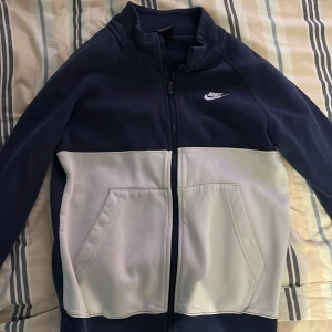 Blå och vit jacka från Nike - Säljer en snygg blå och vit jacka från Nike i mycket bra skick. Den har en dragkedja framtill och två praktiska fickor. Perfekt för höst och vår, med en sportig stil som funkar både till träning och vardag. Jackan är långärmad och gjord i ett mjukt syntetmaterial.