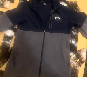 Under Armour jacka i blått och grått - Säljer en snygg och sportig jacka från Under Armour i blått och grått. Jackan har en dragkedja framtill och är perfekt för träning eller vardagsbruk. Den är långärmad och har en bekväm passform. Perfekt för höst och vår!