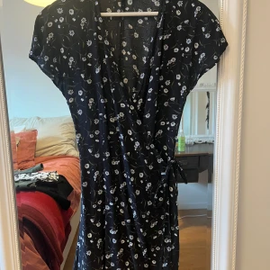 Svart blommig omlottklänning - Säljer en superfin svart omlottklänning med vita blommor. Den har korta ärmar och ett knytband i midjan för en smickrande passform. Perfekt för både vardag och festliga tillfällen!