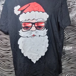 Svart t-shirt med julmotiv - Cool svart t-shirt med ett stort tryck av en jultomte som bär solglasögon med en strandreflektion. Perfekt för att sprida julstämning med en twist! T-shirten är kortärmad och gjord i mjukt material. Perfekt för både jul och sommarvibbar.
