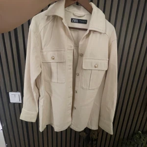 Beige overshirt från Zara - Säljer en snygg beige overshirt från Zara. Den har långa ärmar och två stora fickor framtill med guldknappar. Perfekt för lager-på-lager-stilen under hösten eller våren. Materialet känns mjukt och bekvämt, och den har en klassisk krage. Passar både till vardags och lite mer uppklädda tillfällen. 