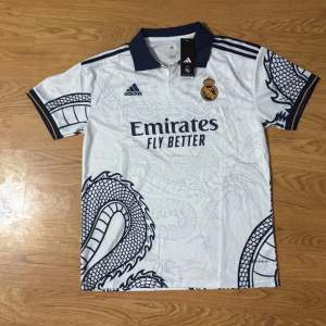 Säljer en vit Real Madrid fotbollströja från Adidas i storlek L. Tröjan har ett coolt drakmönster i grått och klubbens emblem på bröstet. Den är kortärmad med en mörkblå krage och ärmslut. Perfekt för alla fans som vill sticka ut på läktaren! 🐉⚽priset går att diskutera 