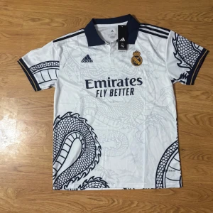 Vit Real Madrid fotbollströja med drakmönster - Säljer en vit Real Madrid fotbollströja från Adidas i storlek L. Tröjan har ett coolt drakmönster i grått och klubbens emblem på bröstet. Den är kortärmad med en mörkblå krage och ärmslut. Perfekt för alla fans som vill sticka ut på läktaren! 🐉⚽priset går att diskutera 