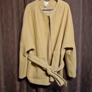 Säljer en stilren beige kappa i mycket bra skick. Den har en enkel design med ett knytband i midjan och långa ärmar. Perfekt för höst och vår när du vill ha något lätt men ändå elegant. Passar både till vardags och lite finare tillfällen.