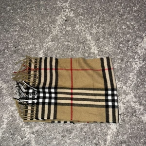 Burberry halsduk  - Säljer en superfin beige halsduk med klassiskt rutigt mönster i svart, vitt och rött. Den är perfekt för att hålla sig varm under höst och vinter. Halsduken har fransar i ändarna som ger en extra touch av stil. Passar både till vardags och festliga tillfällen! Burberry halsduk pris kan diskuteras 