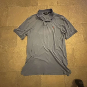 Blå pikétröja från Polo Golf - Säljer en snygg blå pikétröja från Polo Golf i storlek S. Tröjan har en klassisk krage och korta ärmar, perfekt för golf eller vardagsbruk. Den är i bra skick och har en broderad logga på bröstet. Materialet är en blandning av bomull och elastan för extra komfort.