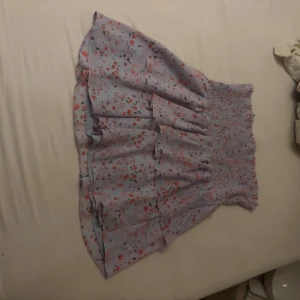 Blommig kortkjol med volang - Superfin blommig kortkjol i ljuslila med volanger. Perfekt för sommardagar eller en mysig vårutflykt. Den har en elastisk midja som gör den bekväm och lätt att bära. Blommönstret i rosa och rött ger en söt och romantisk känsla.