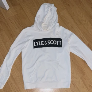 Vit hoodie från Lyle & Scott - Säljer en snygg vit hoodie från Lyle & Scott. Den har en stor logga på bröstet och en klassisk känguruficka. Perfekt för en avslappnad stil och passar till alla årstider. Skön och stilren design!