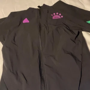 Svart träningsjacka med klubbmärket FC Bayern München från Adidas - Säljer en svart träningsjacka från Adidas med dragkedja och coola detaljer i lila och grönt. Perfekt för träning eller vardagsbruk. Jackan har långa ärmar och är i mycket bra skick där den bara har använts fåtal gånger. Passar perfekt för höst och vår! Ordinarie pris 1136kr