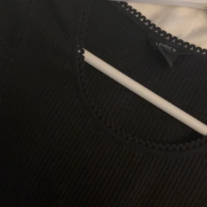Svart ribbad topp från Lindex - Säljer en svart ribbad topp från Lindex i mycket bra skick. Den har en fin spetsdetalj vid halsringningen och ärmarna. Perfekt för både vardag och fest! Passar bra till jeans eller kjol.
