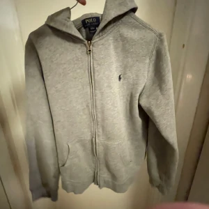 Grå hoodie från Polo Ralph Lauren - Säljer en snygg grå hoodie från Polo Ralph Lauren i bra skick. Den har en dragkedja framtill och en liten broderad logga på bröstet. Perfekt för en avslappnad stil och passar till alla årstider. Storleken är inte angiven men den ser ut att vara normal passform. Det står L men den sitter som S