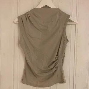Beige ärmlös topp - Snygg beige ärmlös topp i ett mjukt och stretchigt material. Perfekt för sommardagar eller som ett lager under en jacka. Toppen har en enkel design med en lätt rynkad detalj på sidan för en smickrande passform.