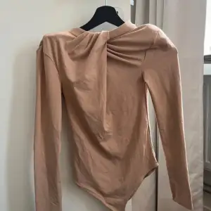 Säljer en elegant beige långärmad topp med en snygg draperad detalj vid halsen. Perfekt för både vardag och festliga tillfällen. Toppen har en tight passform och är gjord i ett mjukt och bekvämt material.