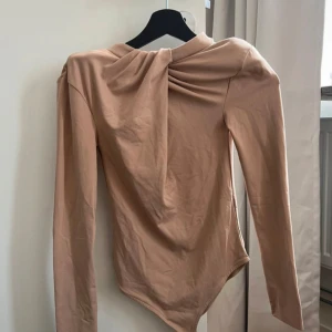 Beige långärmad topp med draperad detalj - Säljer en elegant beige långärmad topp med en snygg draperad detalj vid halsen. Perfekt för både vardag och festliga tillfällen. Toppen har en tight passform och är gjord i ett mjukt och bekvämt material.