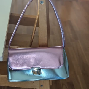 Metallic handväska från Glamorous - Säljer en supercool metallic handväska från Glamorous! Den har en snygg mix av rosa och silver med en stilren design. Väskan har ett praktiskt spänne framtill och en bekväm axelrem. Det finns märken på användningen (vänligen se bilder)