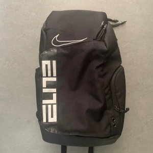 Svart ryggsäck från Nike - Säljer en snygg och praktisk svart ryggsäck från Nike. Den har en stilren design med vit Nike-logga och texten 'ELITE' på framsidan. Ryggsäcken är perfekt för träning eller vardagsbruk med justerbara axelremmar och flera fack för förvaring. Materialet känns slitstarkt och den har en dragkedja för säker förvaring. Perfekt för dig som vill ha en sportig och funktionell väska!