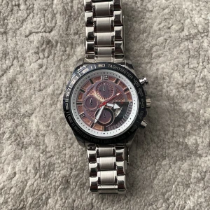 Silverfärgad klocka med tachymeter - Säljer en snygg silverfärgad klocka med tachymeter runt urtavlan. Klockan har en robust metallarmband och en detaljerad urtavla med flera små urtavlor inuti. Perfekt för både vardag och fest! Extra länkar medföljer för justering av storleken.