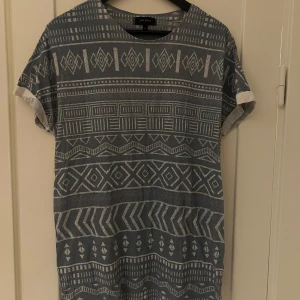 Mönstrad t-shirt från River Island - Säljer en snygg grå t-shirt från River Island med ett coolt geometriskt mönster i vitt. Perfekt för en avslappnad stil, den är kortärmad och gjord i ett mjukt material. Passar perfekt till både jeans och shorts!
