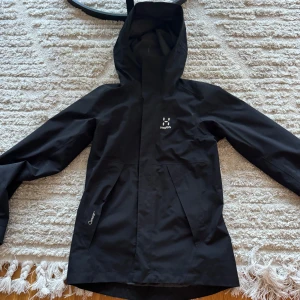 Svart regnjacka från Haglöfs - Säljer en svart regnjacka från Haglöfs i storlek XS för dam. Jackan har en praktisk huva och är perfekt för regniga dagar. Den är tillverkad i ett slitstarkt material och har långa ärmar. Perfekt för höst och vår!