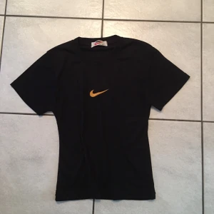 Svart t-shirt från Nike - Snygg svart t-shirt från Nike med det klassiska gula swoosh-loggan på bröstet. Perfekt för en sportig look eller avslappnad stil. Kortärmad och tillverkad i mjukt material för extra komfort.