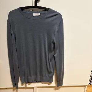 Grå tröja från Tiger of Sweden - Snygg och stilren grå/blå tröja från Tiger of Sweden. Tröjan har en klassisk rund hals och långa ärmar, perfekt för en elegant vardagslook. Materialet är mjukt och bekvämt, vilket gör den idealisk för både höst och vår.