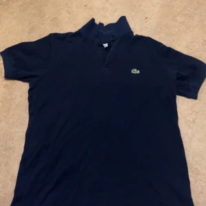 Mörkblå pikétröja från Lacoste - Snygg mörkblå pikétröja från Lacoste med klassisk krage och knappslå. 