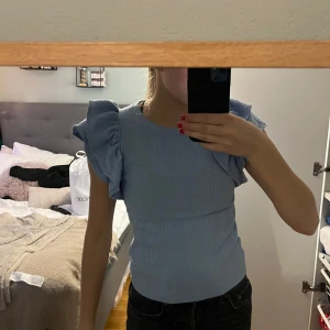 Ljusblå ribbad topp med volangärmar - Säljer en söt ljusblå ribbad topp med volangdetaljer på ärmarna. Perfekt för en stilren och trendig look. Toppen har en tight passform och ärmlös design.