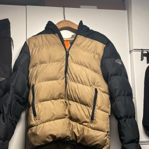 Beige och svart pufferjacka från Replay - Snygg pufferjacka från Replay i beige och svart. Jackan har en dragkedja framtill och två praktiska fickor med dragkedjor. Den har en huva och är perfekt för kyligare dagar. Märkeslogga på ärmen ger en extra touch. Hör av er vid frågor