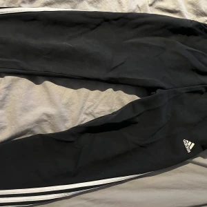 Svarta träningsbyxor från Adidas - Säljer ett par svarta träningsbyxor från Adidas med vita ränder längs sidorna. Byxorna har en bekväm passform och är perfekta för träning eller avslappning. Adidas-loggan är synlig på ena benet.