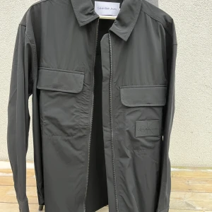 overshirt från Calvin Klein Jeans - Snygg svart overshirt från Calvin Klein Jeans med dragkedja och två stora bröstfickor. Perfekt för lager-på-lager-stil. Märkeslogga på bröstet ger en stilren touch. Aldrig använt så den är som ny.