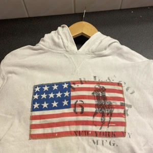 Vit hoodie från Polo Ralph Lauren - Säljer en vit hoodie från Polo Ralph Lauren med ett stort amerikanskt flaggmotiv och en hästtryck på framsidan. Tröjan har en klassisk huva och är perfekt för en avslappnad stil.