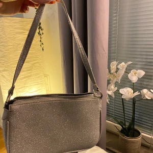 Glittrig silver axelväska - Snygg glittrig axelväska i silver med dragkedja. Perfekt för att ge en extra touch till din outfit. Väskan har en praktisk storlek och en bekväm axelrem.
