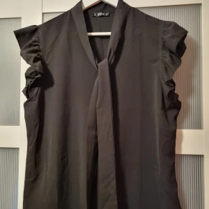 Svart blus med volangdetaljer - Snygg svart ärmlös topp med volangdetaljer vid ärmhålen. Perfekt för en stilren look. Toppen har en enkel och elegant design som passar till många olika tillfällen.