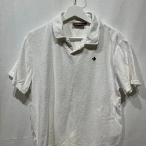 Vit frotté piké från Morris - Säljer en stilren vit t-shirt från Morris i frotté med krage och korta ärmar. Den har en liten broderad detalj på bröstet. Perfekt för en avslappnad och snygg look. Modellen är 180cm