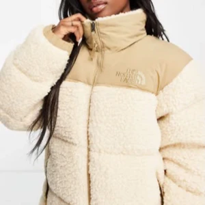 North Face jacka - Mysig beige teddyjacka från The North Face med hög krage och dragkedja. Jackan har en pufferdesign och är perfekt för kyliga dagar. Den har långa ärmar och en kontrasterande beige panel upptill med logotypen på bröstet.