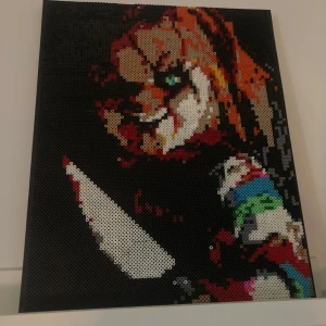 Pärlplatta med skräckmotiv, chucky - En pärlplatta med ett skräckmotiv i olika färger, inklusive svart, rött, vitt och grönt. Motivet föreställer en figur med en kniv, vilket ger en dramatisk och spännande känsla. Perfekt för den som gillar skräck och unika konstverk.                                                               50 x40 cm.
