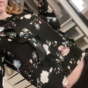 Svart blommig blus med volangdetaljer - Snygg svart blus med blommigt mönster i vitt, blått och rosa. Blusen har långa ärmar och volangdetaljer framtill som ger en romantisk touch. Perfekt för att piffa upp vardagslooken eller för en speciell kväll.