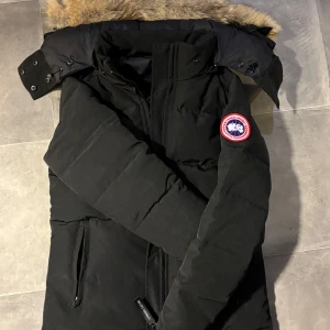 Svart dunjacka från Canada Goose - Säljer en svart dunjacka från Canada Goose med pälsfodrad huva. Jackan har en broderad logotyp på ärmen och dragkedja framtill. Perfekt för kalla vinterdagar.