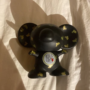 Svart Koala Bay figur - Unik svart figur från Koala Bay med små gula koalor som dekoration. Perfekt som samlarobjekt eller dekoration i hemmet. Figuren har stora runda öron och en rund kropp med logotypen tydligt synlig framtill. 💕