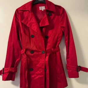 Röd trenchcoat från Stockh LM - Snygg röd trenchcoat från Stockh LM i storlek 34. Jackan har knappar framtill och ett bälte i midjan för en figursydd look. Perfekt för höst och vår med sina långa ärmar och klassiska design.